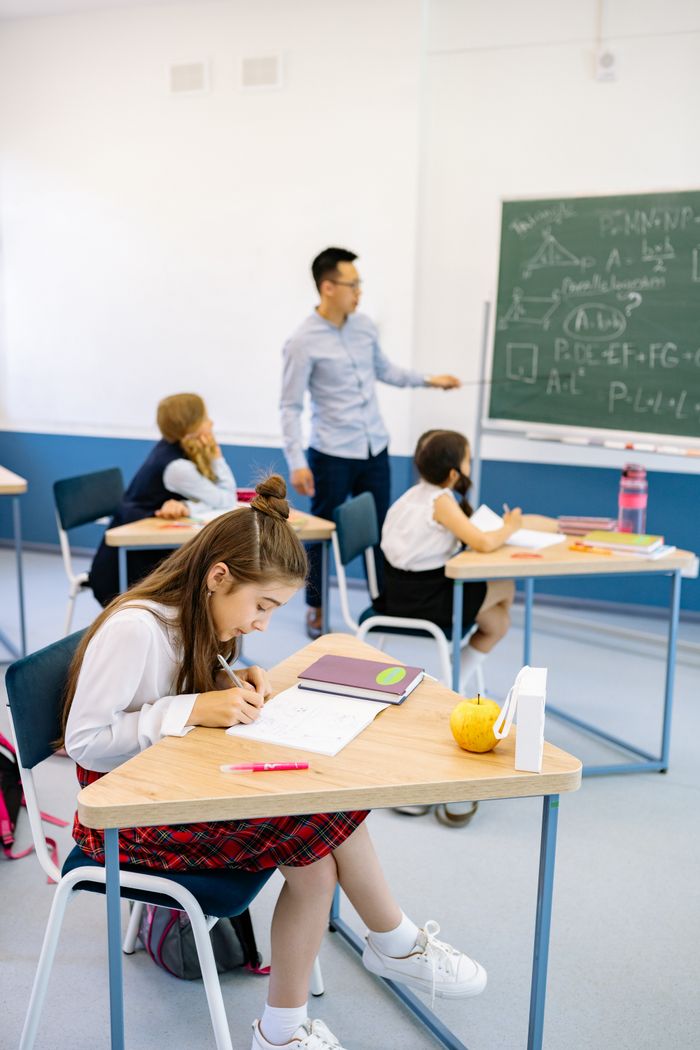 A teacher stands near a blackboard teaching students seated at desks in a classroom.